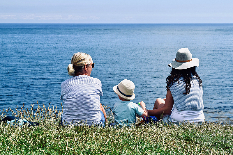 Two women and a child sit on a cliff overlooking the ocean