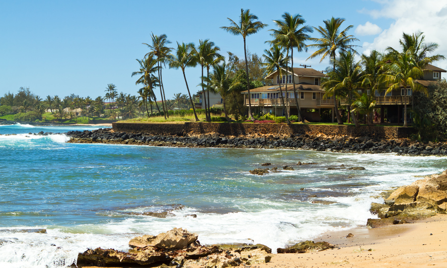 House surrounded by tall palm trees facing the beautiful blue ocean and a sandy beach at south shore of Kauai, Hawaii on a gorgeous sunny, summer day in the tropics