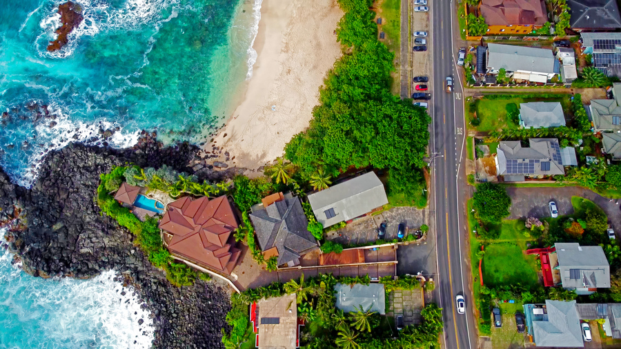 Aerial View of Luxury Beachfront Homes in Hawaii. A breathtaking aerial shot of beachfront houses along a tropical coastline in Hawaii, featuring turquoise ocean waves, sandy beach, and lush greenery